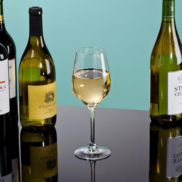 A close-up of a Arcoroc wine glass filled with white wine next to bottles of wine.