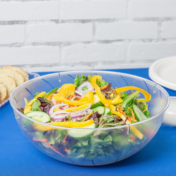 A salad in a clear Fineline plastic bowl on a table with bread and other food.