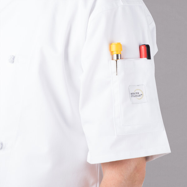 A person wearing a white Mercer Culinary chef jacket with a pocket full of pens.