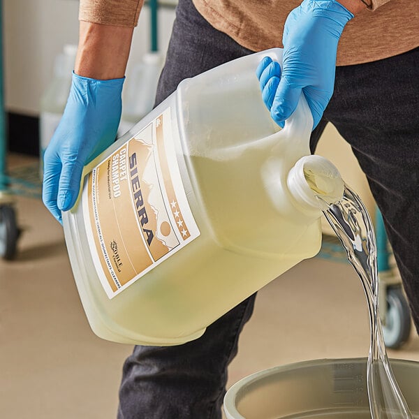 A person in blue gloves pouring Sierra by Noble Chemical concentrated carpet shampoo into a bucket.