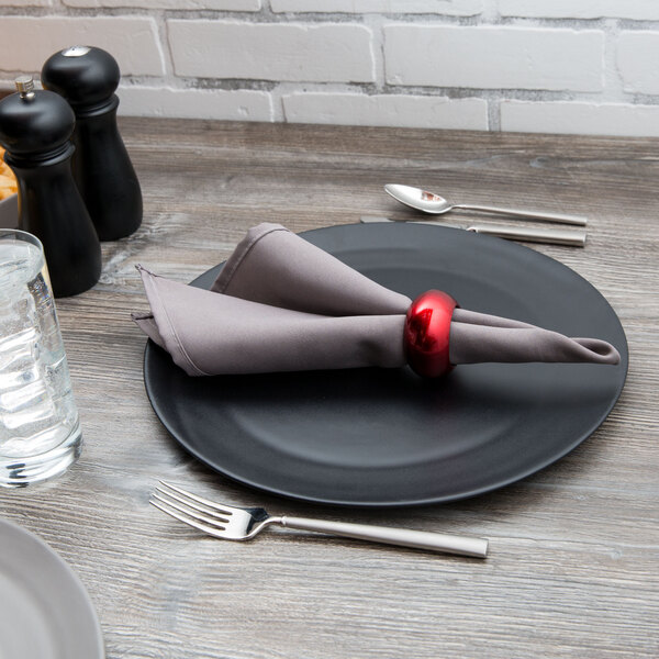 A black 10 Strawberry Street stoneware charger plate with a napkin and fork on a table.