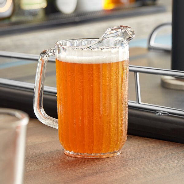 A Cambro clear plastic pitcher on a counter with a glass of orange juice in it.