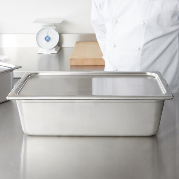 A chef in a white coat standing behind a Vollrath stainless steel pan cover on a metal counter.