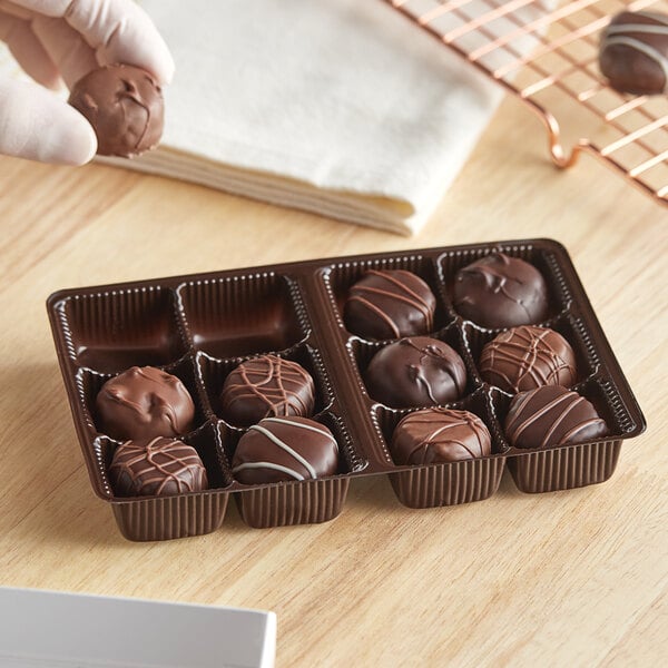 A hand holding a chocolate truffle in a brown plastic candy tray.