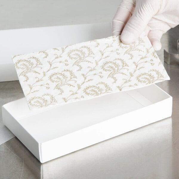 A gloved hand holding a white glassine pad with a gold floral pattern.