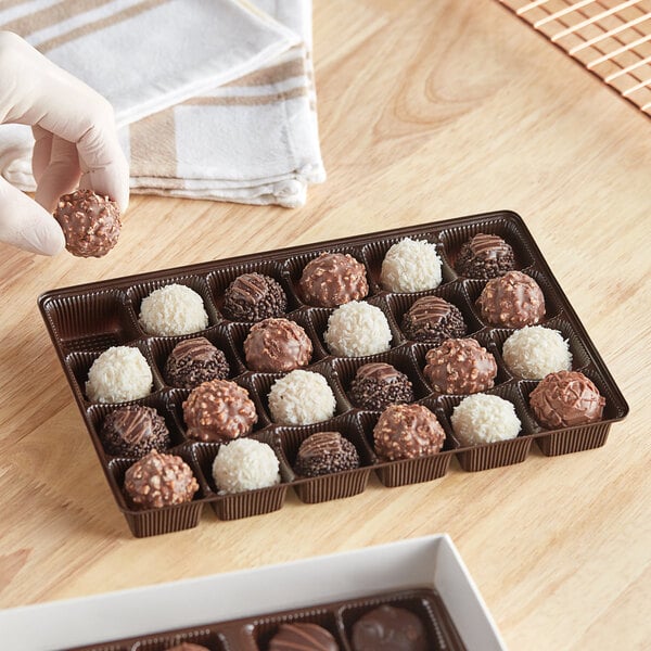 A hand holding a chocolate ball in a brown candy tray.