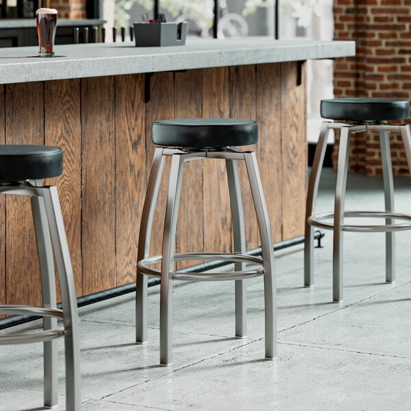 A row of Lancaster Table & Seating black vinyl bar stools in front of a bar.