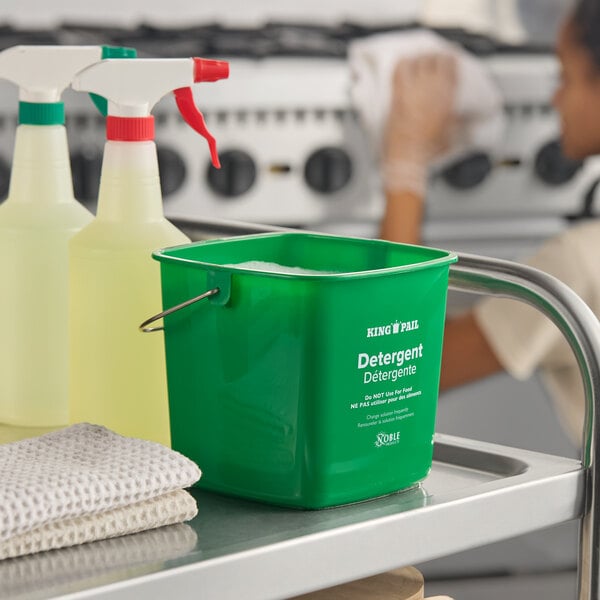 A green Noble King-Pail 3 quart cleaning pail placed on a metal cart next to spray bottles and cleaning cloths.