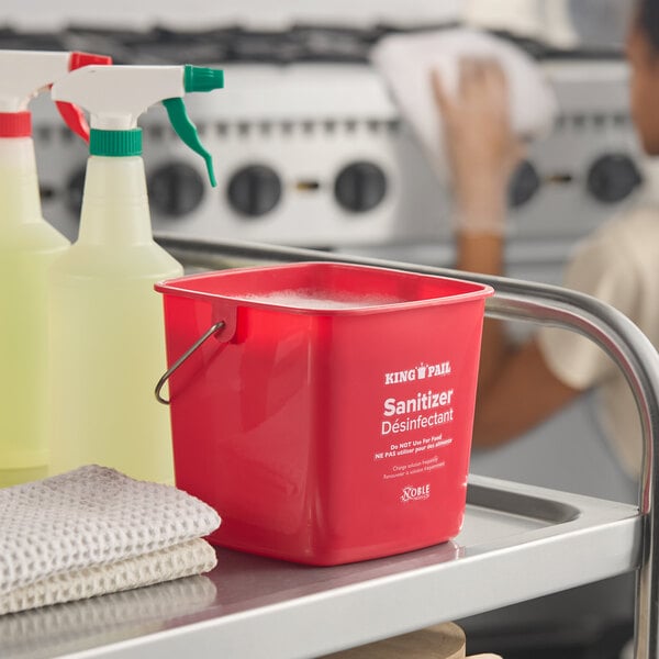 A red 3-quart sanitizing pail labeled 'Sanitizer Désinfectant' by Noble King-Pail, placed on a cleaning cart with spray bottles and cloths.