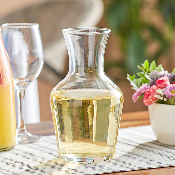 A glass wine carafe filled with yellow liquid on a table with a glass of wine.