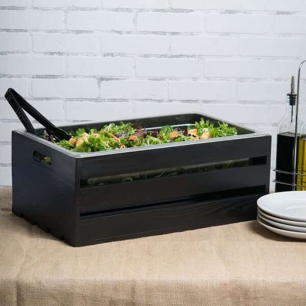 A wooden American Metalcraft crate with a salad inside on a table with white plates and tongs.