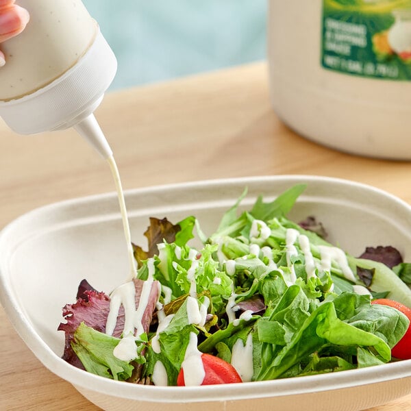 A fresh salad in a disposable bowl being drizzled with ranch dressing from a squeeze bottle.