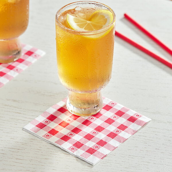 A glass of liquid with a lemon slice and a red and white checkered napkin with two straws.