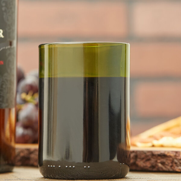 An Arcoroc green wine bottle tumbler filled with wine on a table next to a bottle of wine.