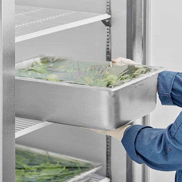 A person in blue gloves holding a Vollrath stainless steel steam table pan of food opening a refrigerator.