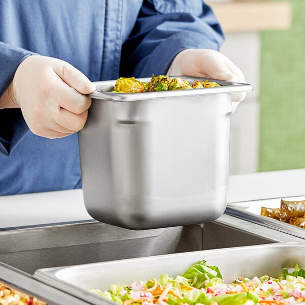 A person in a blue uniform holding a Vollrath stainless steel container of food.