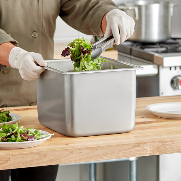 A person in gloves using tongs to fill a Vollrath stainless steel container with salad at a salad bar.