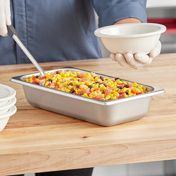 A person holding a stainless steel hotel pan filled with food and a spoon.