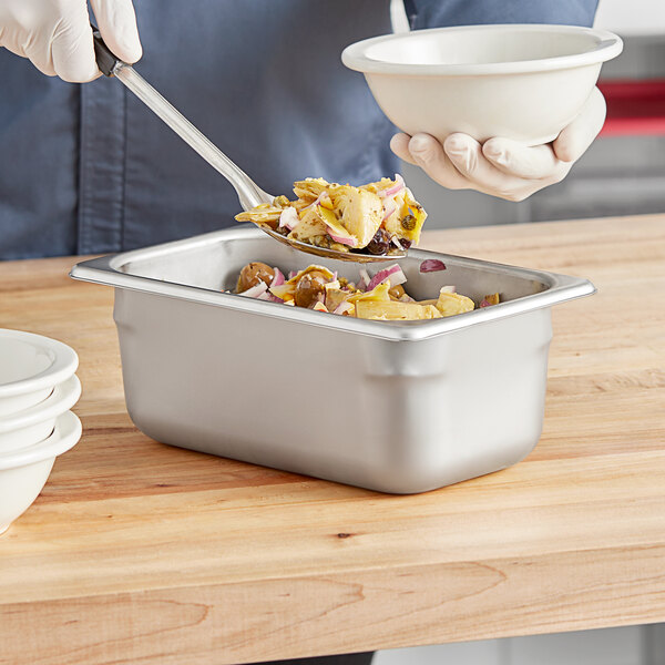 A silver Vollrath steam table pan filled with food on a counter with a white bowl and spoon.