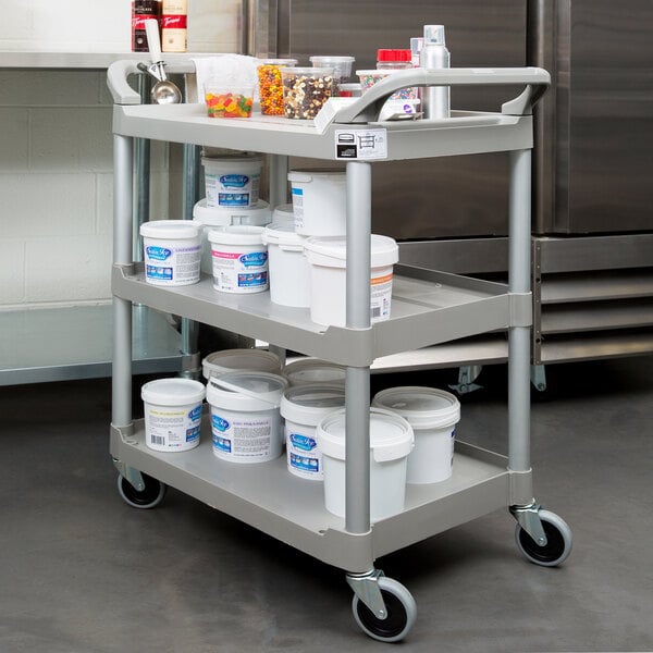 A Rubbermaid platinum three shelf utility cart with white food containers on it.