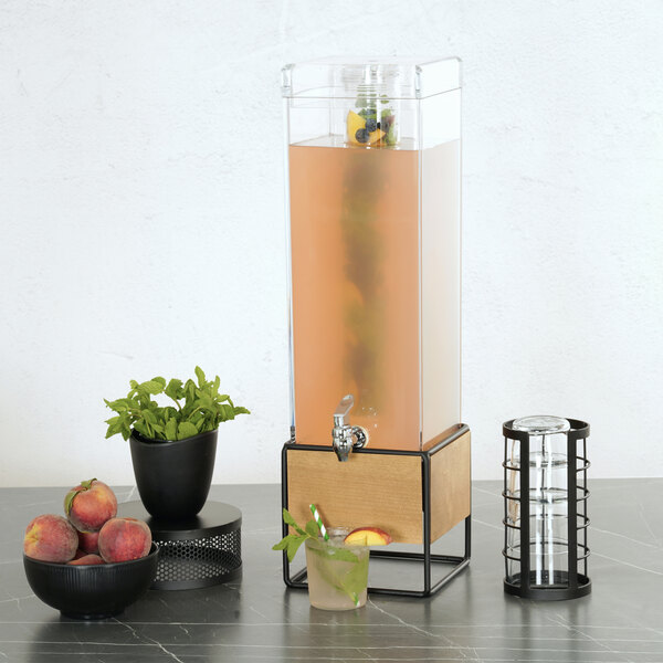 A square 3-gallon beverage dispenser with a walnut and metal base and an infusion chamber, shown on a countertop with fruit, mint, and glasses nearby.