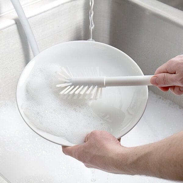 A hand holding a Carlisle Sparta dish brush over a bowl of soapy water.