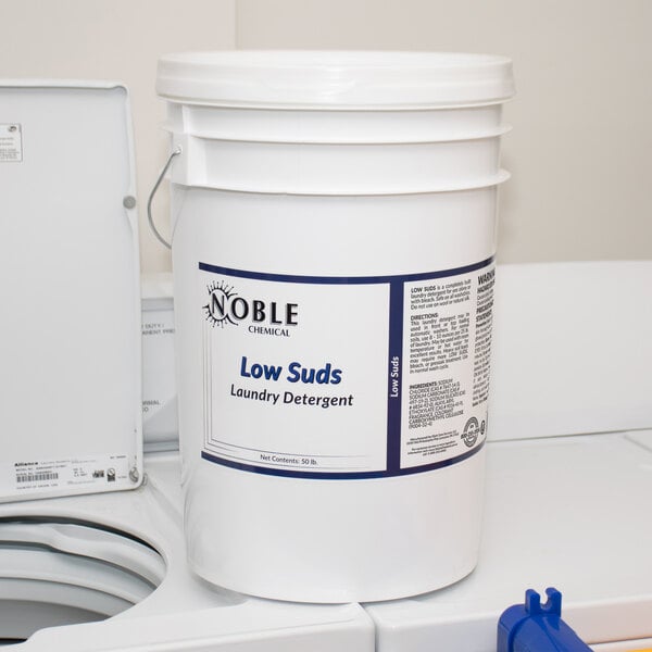 A white bucket of Noble Chemical laundry detergent powder with a black handle on a white counter.