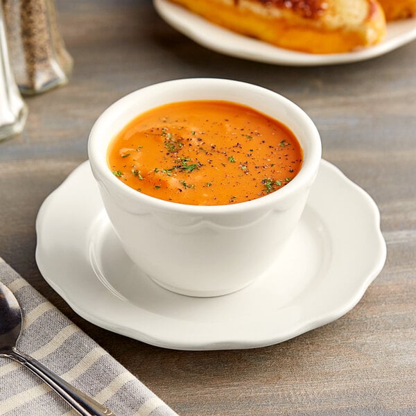 An Acopa ivory stoneware bouillon cup filled with soup on a table with bread and a spoon.