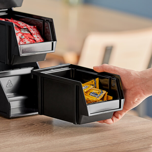 A hand holding a Vollrath black self-serve condiment bin with red and yellow packets inside.