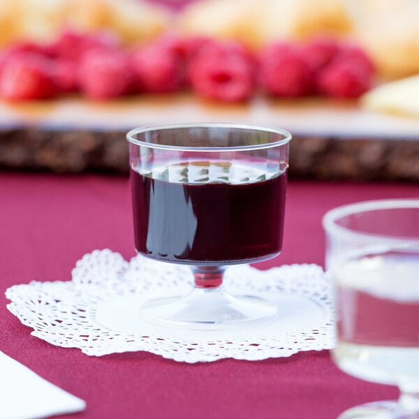 A WNA Comet Classicware clear plastic pedestal wine cup filled with red liquid on a table with a doily.