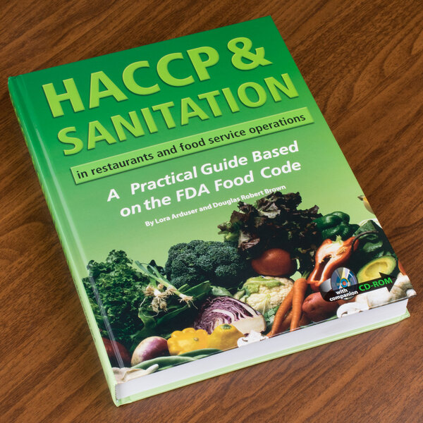 A green HACCP & Sanitation book on a wood table near a variety of vegetables.