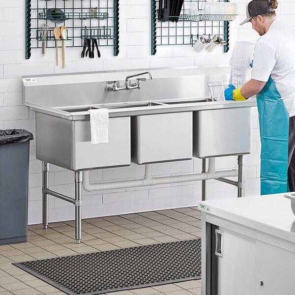 A man washing dishes in a commercial kitchen using a Regency three compartment sink.