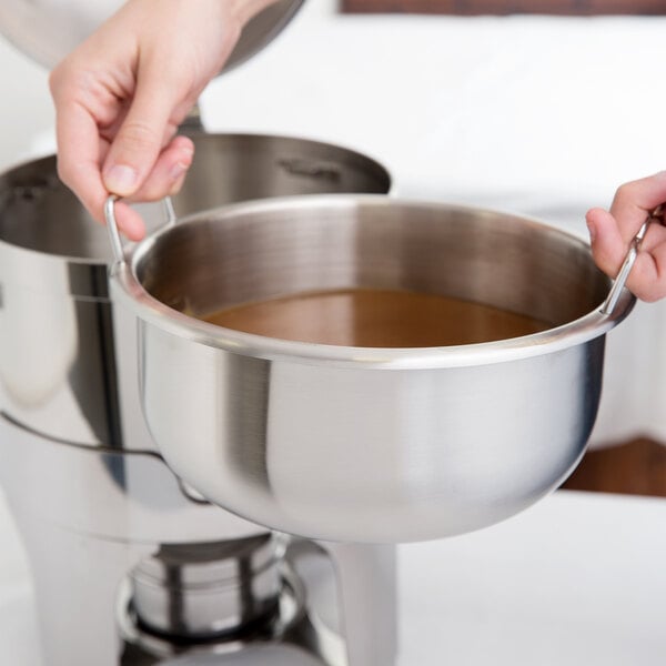 A hand using a Vollrath stainless steel inset to pour brown liquid into a silver pot.