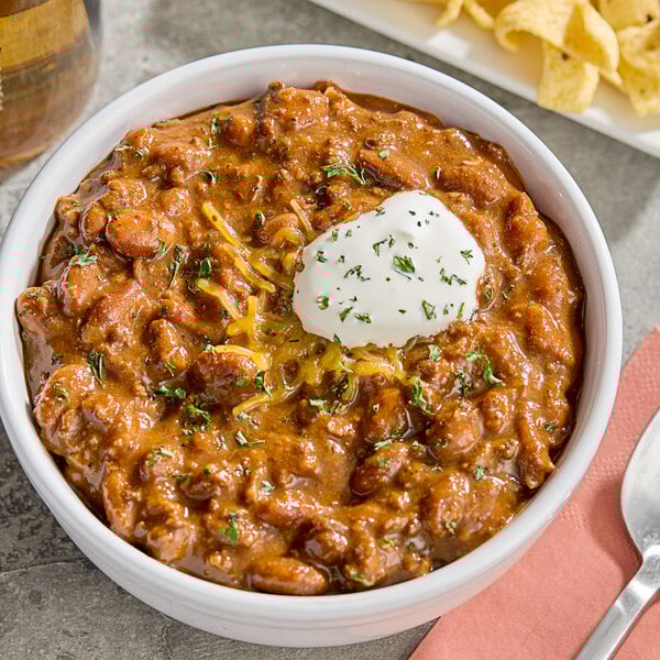 A bowl of Vanee chili with beans with a spoon and a plate of chips.