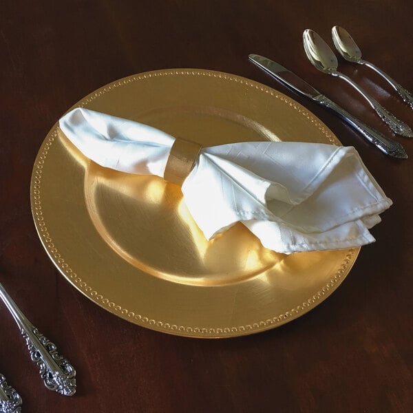 A Tabletop Classics gold charger plate with a napkin and silverware on a table.