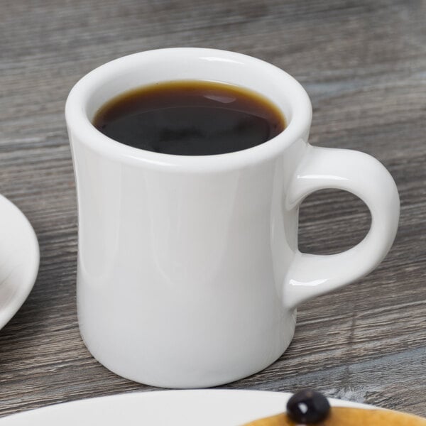 A Tuxton white china mug filled with a brown liquid on a white surface.