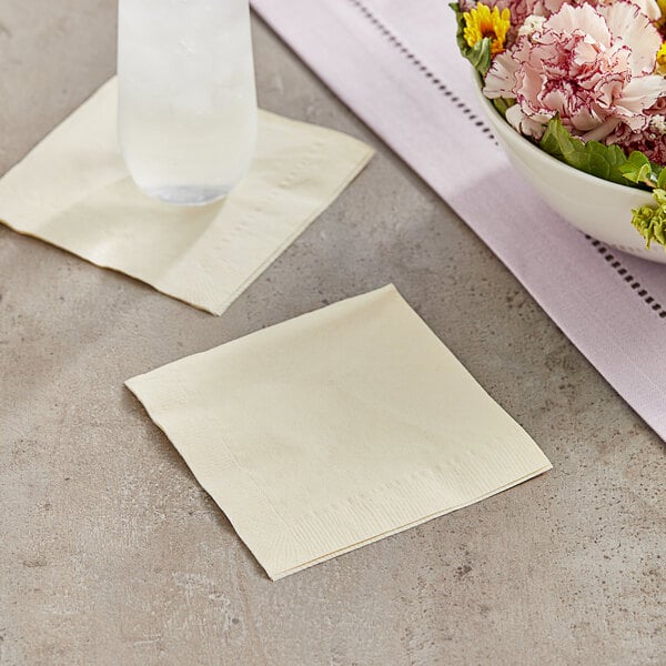A Choice ivory 2-ply customizable beverage napkin next to a glass of water on a table with a bowl of flowers.