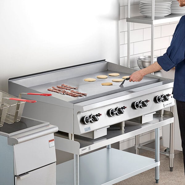 A large stainless steel 48-inch liquid propane countertop griddle with thermostatic controls, shown cooking bacon and pancakes in a commercial kitchen setting.