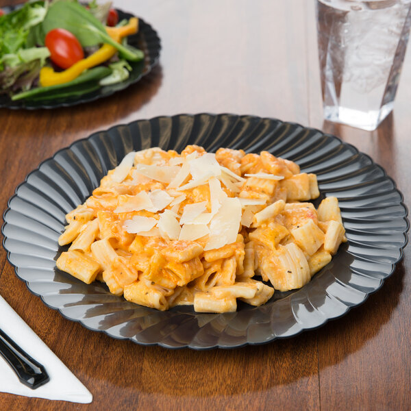 Pasta on a scalloped black biodegradable plate with a salad in the background on a matching plate