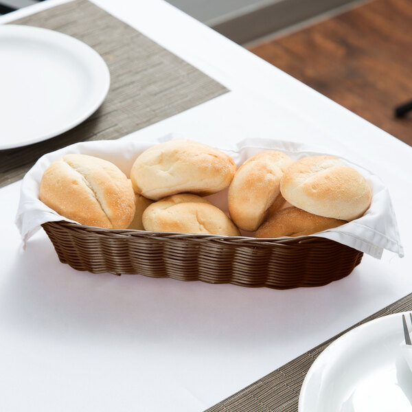 A Tablecraft brown rattan bread basket filled with rolls on a table.