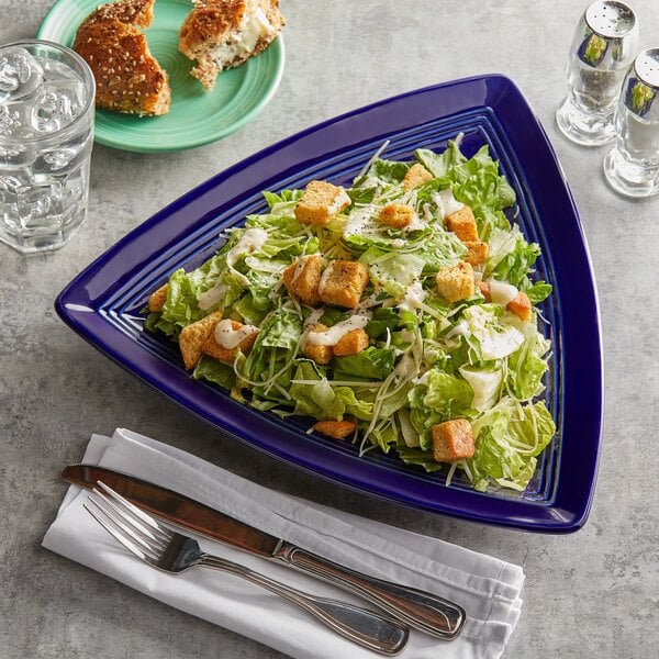 A blue Tuxton Concentrix triangle china plate with a salad on it on a table next to other food.