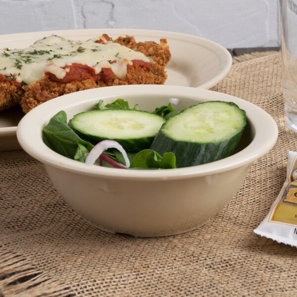 A Carlisle tan melamine nappie bowl filled with cucumbers and lettuce on a table with a bowl of salad.