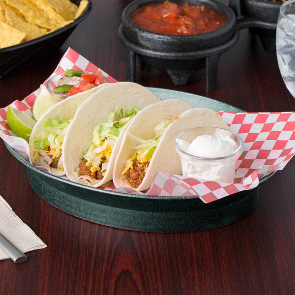 A plate of tacos and a bowl of chips and salsa served in a red and white checkered oval deli server.