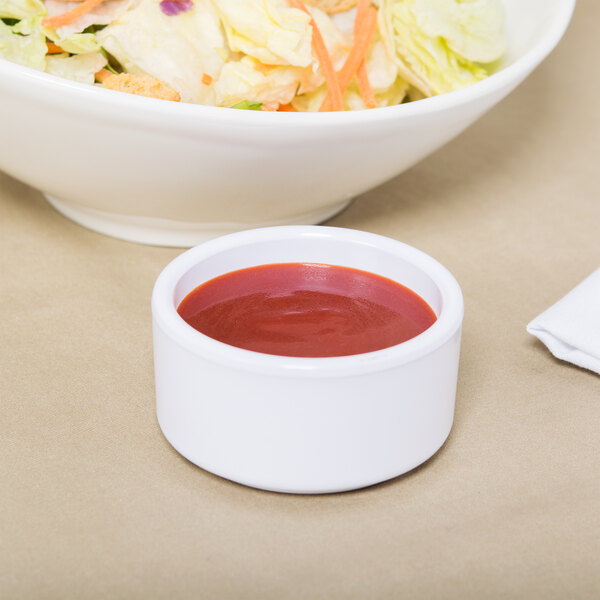 A white Carlisle ramekin filled with red sauce next to a bowl of salad with vegetables.