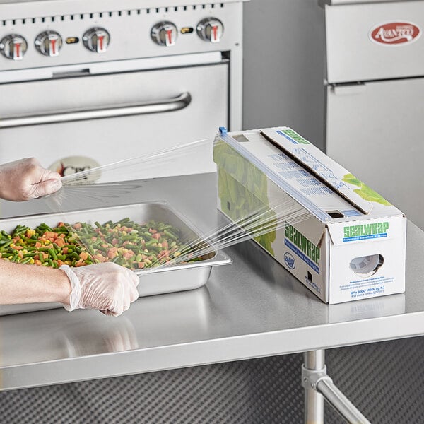 A person in gloves and apron using Berry heavy-duty plastic wrap to cover a tray of vegetables.