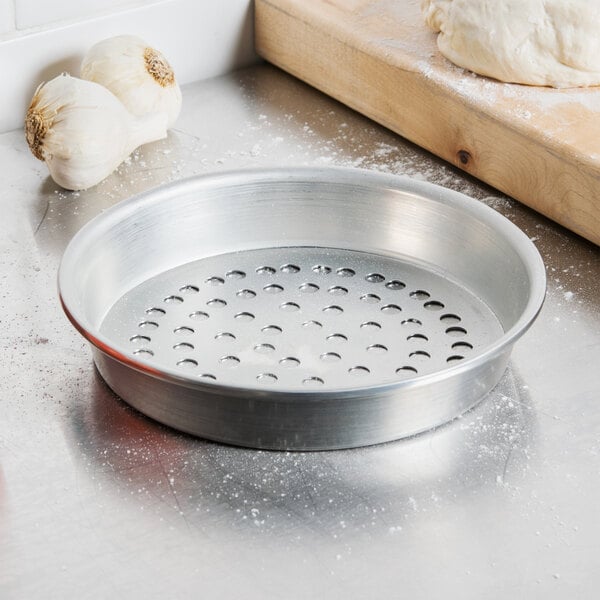 An American Metalcraft tapered pizza pan with holes on it next to a wooden cutting board and garlic.