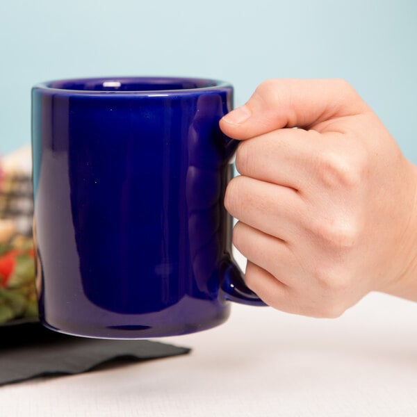 A hand holding a Tuxton cobalt blue C-handle mug.