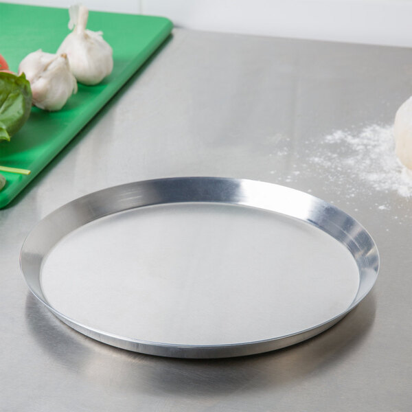 An American Metalcraft aluminum pizza pan with a white surface on a counter next to vegetables.