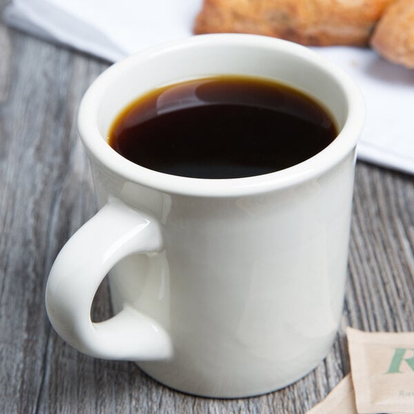 A white Tuxton china coffee mug filled with brown liquid on a table with a plate of pastries.