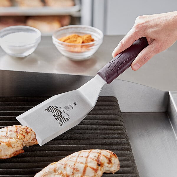 A hand using a Mercer Culinary Hell's Handle spatula to cook chicken on a grill.